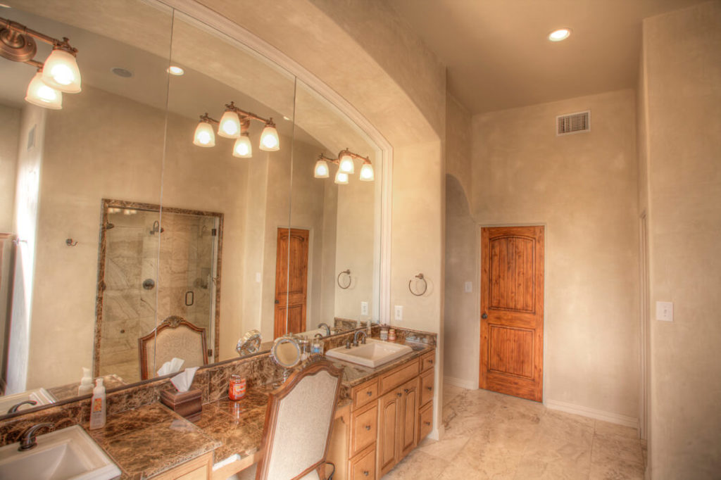 Primary bathroom with large mirrors and makeup vanity. Wood-grain cabinets, double sinks, high ceilings, and tan tile floor.